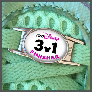 Può includere: Un ciondolo per scarpe argentato con un cerchio bianco che presenta il testo "runDisney 3.1 FINISHER" in lettere nere e rosa. Una testa di Topolino è al centro del numero "3.1".