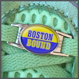 May include: A silver shoelace charm with a yellow, blue and white oval design. The oval has the text "BOSTON BOUND" in white and yellow letters.