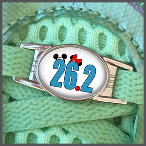 Può includere: Un ciondolo per lacci delle scarpe argentato con sfondo bianco e un numero blu "26.2" con un punto rosso. Topolino e Minnie Mouse sono in cima al ciondolo.
