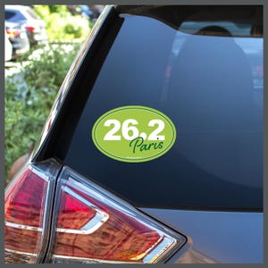 May include: A green oval sticker with the text "26.2 Paris" in white and green lettering is affixed to a car window. The sticker is on the rear window of a gray car, near the taillight.