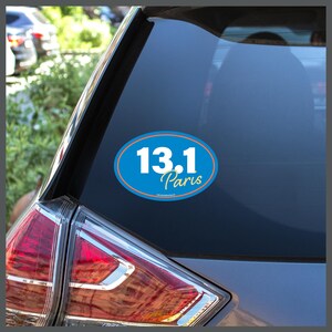 May include: An oval sticker with the text "13.1 Paris" in white and gold lettering, set against a blue background. The sticker is affixed to the rear window of a gray vehicle, near the taillight.