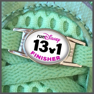 Può includere: Un ciondolo per scarpe argentato con un'etichetta ovale bianca che dice "runDisney 13.1 FINISHER" in lettere nere e rosa. Il ciondolo ha una testa di Topolino accanto al numero 1.