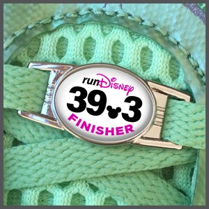 Può includere: Un ciondolo per scarpe argentato con un'etichetta ovale bianca che dice "runDisney 39.3 FINISHER" in lettere nere e rosa. Il ciondolo ha una testa di Topolino al centro del numero 3.