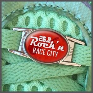 Peut inclure: Gros plan sur un lacet de chaussure vert clair avec un fermoir argenté portant l'inscription "26.2 Rock'n Race City" en blanc sur un ovale rouge. Le fond est un tissu en maille vert clair.