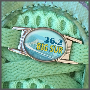 以下が含まれることがあります： 黄色と青で「26.2 BIG SUR」の文字が入った、銀色の靴紐チャーム。風景を背景に、明るい緑色の靴紐の上に配置されています。背景には、お揃いの緑色のニット生地があります。