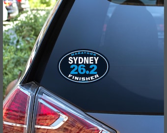 May include: An oval sticker on a car window. The sticker has a dark blue background with white text that reads "MARATHON SYDNEY 26.2 FINISHER" in a circular design. The car's taillight is visible.