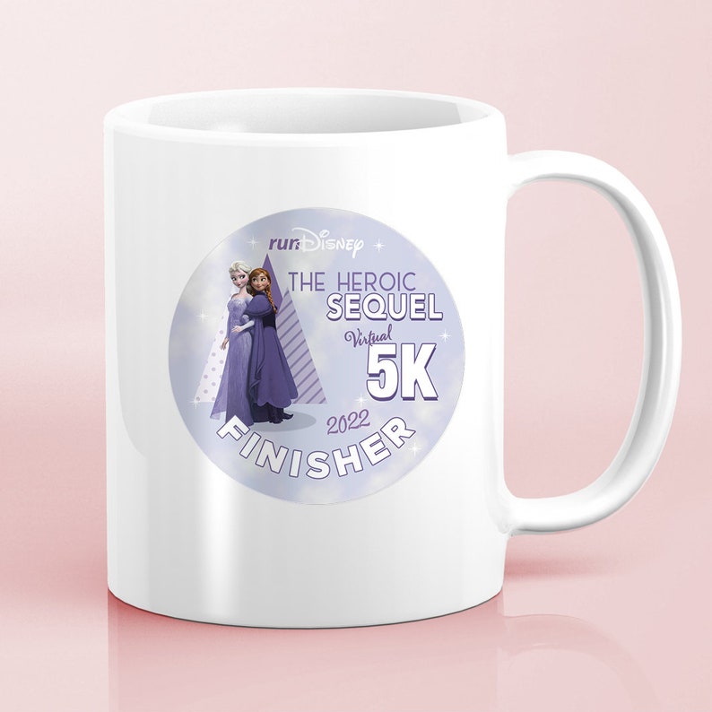 May include: White ceramic mug with a purple and blue circular sticker featuring the text "runDisney The Heroic Sequel Virtual 5K 2022 Finisher" and an image of two cartoon women.