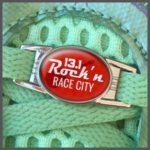 Peut inclure: Gros plan sur un fermoir de lacet argenté avec un ovale rouge portant l'inscription "13.1 Rock'n Race City". Le fermoir est fixé à un lacet vert clair, sur un fond en maille verte assortie.