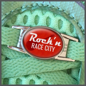 Peut inclure: Un charme de lacet de chaussure argenté avec une étiquette circulaire rouge qui dit "Rock'n RACE CITY" en texte blanc. L'étiquette a un graphique de guitare.