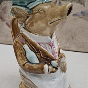 Può includere: Brocca in ceramica a forma di maiale, con cappello e giacca da chef. La brocca è principalmente marrone con un grembiule bianco e un cappello verde-blu. Ha un beccuccio per versare e un manico laterale. Il viso del maiale è dettagliato.