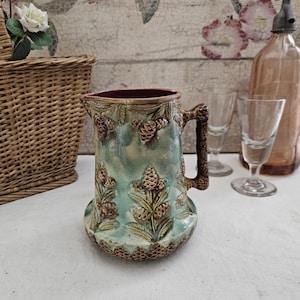 Puede incluir: Una jarra de cerámica vintage con un esmalte turquesa y marrón, decorada con diseños en relieve de piñas y hojas. La jarra tiene un interior rojo oscuro y un asa texturizada. Una pieza decorativa para el hogar.