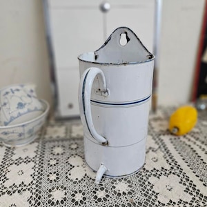 Op de afbeelding: Witte geëmailleerde wandhouder voor keukengerei met een blauw gestreept ontwerp en een handvat. De houder heeft een klein gaatje bovenaan om op te hangen.