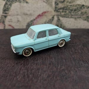 Può includere: Un'auto giocattolo azzurra con quattro porte e ruote bianche. L'auto modello vintage è ambientata su una superficie marrone scuro e uno sfondo a motivi. Il design dell'auto include una griglia anteriore dettagliata e specchietti laterali.