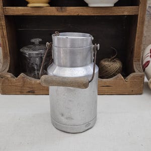 Könnte beinhalten: Eine alte Milchkanne aus Aluminium mit Holzgriff. Die Kanne ist zylindrisch mit Deckel und Metallgriff. Sie steht auf einer weißen Oberfläche, im Hintergrund weitere Küchenartikel, darunter Schalen und ein Regal.