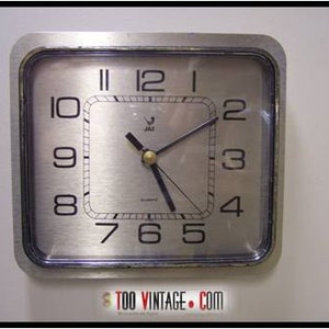 Könnte beinhalten: Eine silberne quadratische Wanduhr mit schwarzen Zeigern und schwarzen Zahlen. Das Zifferblatt der Uhr hat einen silbernen Hintergrund mit einer gebürsteten Metalloberfläche. Die Uhr wird von JAZ hergestellt und trägt die Aufschrift "Quartz" auf dem Zifferblatt.