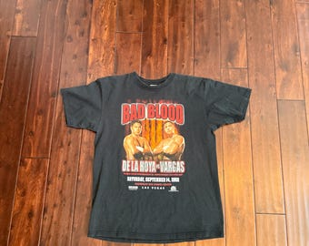 Könnte beinhalten: Schwarzes T-Shirt mit dem roten Text "BAD BLOOD" und einem Bild von zwei Boxern. Darunter stehen die Namen "DE LA HOYA-VARGAS" sowie Veranstaltungsdetails: "SAMSTAG, 14. SEPTEMBER 2002, MANDALAY BAY EVENTS CENTER, LAS VEGAS."