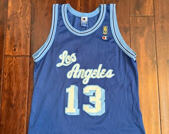 Könnte beinhalten: Blaues Champion Basketballtrikot mit den Worten "Los Angeles" und der Nummer "13" in Weiß und Hellblau. Das Trikot hat hellblaue und weiße Verzierungen. Ein NBA-Logo ist sichtbar. Maße in Zentimetern.