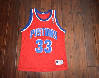 Könnte beinhalten: Rotes Basketballtrikot mit blauem und weißem Besatz. Das Trikot hat "Pistons" in blauen Buchstaben über der Brust und die Nummer "33" in blau darunter. Das Trikot ist von Champion und hat die Größe 40.