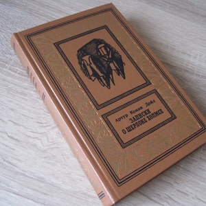 May include: A brown hardcover book with gold lettering on the spine. The title is "The Adventures of Sherlock Holmes" by Arthur Conan Doyle.