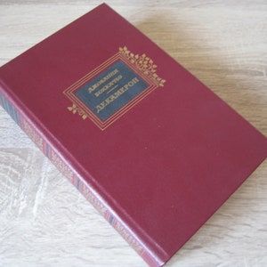 Może przedstawiać: Burgundowa książka w twardej oprawie ze złotymi napisami na grzbiecie i okładce. Tytuł książki to "Giovanni Boccaccio Decameron".