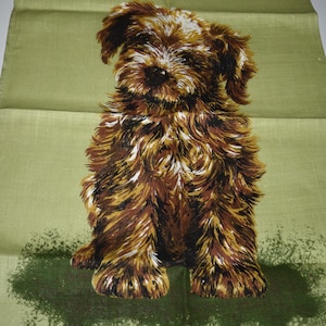 May include: A green tea towel with a brown and white illustration of a puppy. The text "Guide Dogs for the Blind" is printed in black at the top of the towel. The text "IRISH LINEN" is printed in black at the bottom left corner of the towel. The text "SCRUFFY BY ULSTER REG NO 7915" is printed in black at the bottom center of the towel. The text "MADE IN" is printed in black at the bottom right corner of the towel.