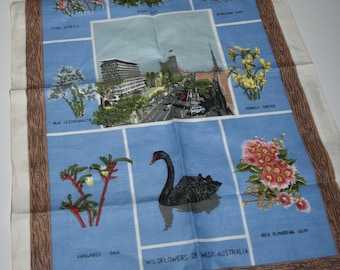 May include: A blue linen tea towel featuring illustrations of West Australian wildflowers and a black swan. The towel includes the text "Wildflowers of West Australia" and "Made in China". The border is a brown wood grain pattern.