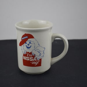 May include: A white ceramic mug featuring a graphic of a cartoon character wearing a red cap with the Nissan logo. The character is pointing forward, and the text below reads "I'm NISSAN-ing!" in red letters. The mug has a curved handle and a smooth finish.