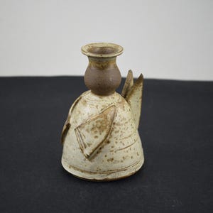May include: A ceramic candle holder shaped like an angel with a speckled cream-coloured body and a solid brown neck and rim. The figure features triangular wings on the sides and a flared base, crafted from textured clay with visible brown spots.