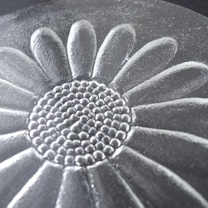 May include: A circular clear glass plate featuring a raised, embossed daisy flower design in the center. The flower has a textured, dotted circular core surrounded by elongated, smooth petals that extend toward the edge of the glass disc.