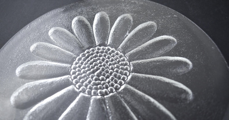 May include: A circular clear glass plate featuring a raised, embossed daisy flower design in the center. The flower has a textured, dotted circular core surrounded by elongated, smooth petals that extend toward the edge of the glass disc.