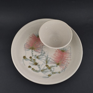 May include: A ceramic plate and bowl set. The off-white plate features a floral design with pink and green accents. The bowl is a matching off-white colour and sits atop the plate. The set is on a dark background.