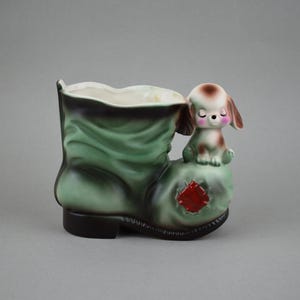 May include: A green ceramic boot planter with a brown and white dog figurine on top. The boot has a red patch on the side.