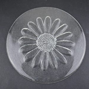 May include: A circular clear glass plate featuring a molded, raised daisy flower design in the center. The flower has a textured, dotted circular core surrounded by numerous elongated, smooth petals that extend toward the plate's edge.