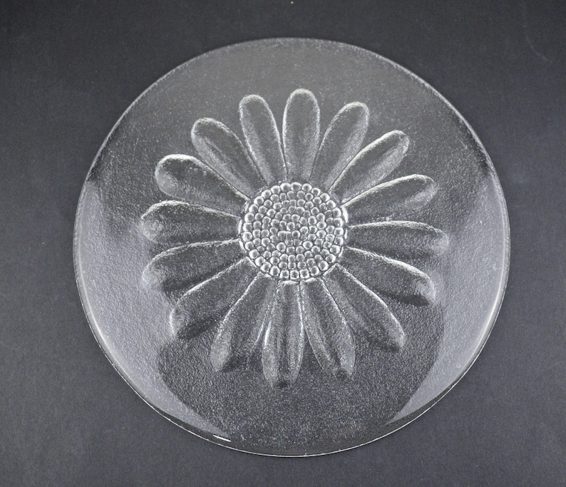 May include: A circular clear glass plate featuring a molded, raised daisy flower design in the center. The flower has a textured, dotted circular core surrounded by numerous elongated, smooth petals that extend toward the plate's edge.
