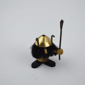 May include: A small wooden figurine of a Viking warrior. The figure has a gold-colored helmet, a black furry beard, and holds a spear. The base and feet are dark brown.