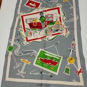 May include: A vintage gray tea towel with a whimsical car-themed design. The towel features illustrations of classic cars, road signs, and musical notes, framed by a white border. Text includes "Oh Genevieve", "Detour", and "Washable Colours".