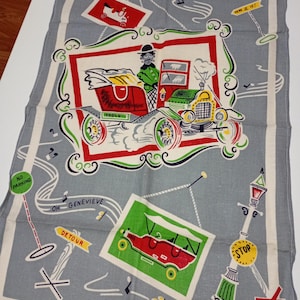 May include: A vintage gray tea towel with a whimsical car-themed design. The towel features a central framed image of a classic car with a driver, surrounded by smaller car illustrations, road signs, and musical notes. The words "Washable Colours" are printed at the bottom.