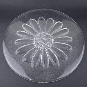 May include: A circular clear glass plate featuring a textured, embossed daisy flower design in the center. The flower has a detailed stippled circular core surrounded by numerous elongated petals that extend outward toward the smooth, rounded rim.