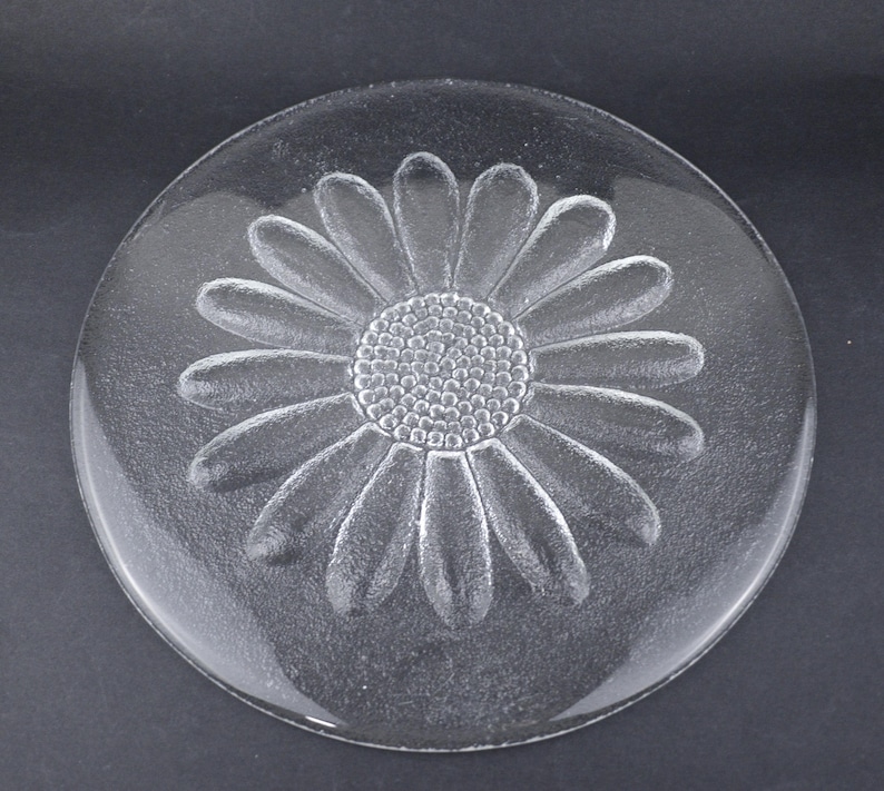 May include: A circular clear glass plate featuring a textured, embossed daisy flower design in the center. The flower has a detailed stippled circular core surrounded by numerous elongated petals that extend outward toward the smooth, rounded rim.
