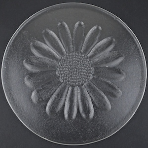 May include: A circular clear glass plate featuring a raised, textured daisy flower design in the center. The petals and the central disc of the flower are embossed, creating a tactile surface on the otherwise smooth and transparent glass material.
