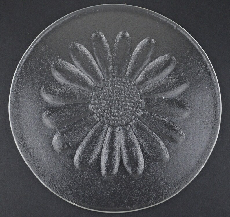 May include: A circular clear glass plate featuring a raised, textured daisy flower design in the center. The petals and the central disc of the flower are embossed, creating a tactile surface on the otherwise smooth and transparent glass material.