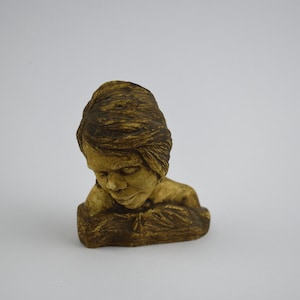 May include: A small, detailed sculpture of a person with their head bowed. The sculpture is a warm, earthy brown color, with visible texture on the hair and face. The figure appears to be resting their head on their hand.