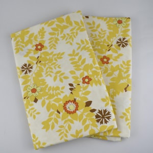 May include: Two folded sheets with a floral pattern. The sheets feature a cream-colored background with a repeating pattern of yellow leaves and flowers in shades of orange, brown, and yellow. The sheets are likely made of cotton or a similar fabric.