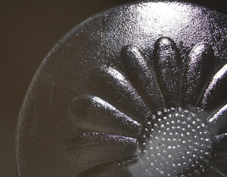 May include: A clear glass circular object featuring a raised, molded daisy flower design. The center of the flower has a textured, dotted surface, while the surrounding petals are smooth and elongated, radiating outward toward the rounded edge.