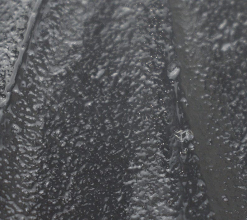 May include: A dark gray, textured surface with a bumpy, uneven finish. The material appears to have a slightly glossy or wet sheen, with small, irregular protrusions scattered across the entire area, creating a tactile and rugged visual appearance.