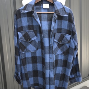 May include: A blue and black checked shirt with two chest pockets, displayed on a wooden hanger and a white mannequin form. The shirt features a classic collar and buttoned cuffs. The fabric appears to be a wool blend.