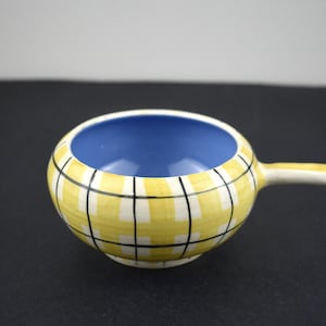 May include: A small, round ceramic bowl with a handle. The exterior is yellow with a black and white grid pattern. The interior is a bright blue. The handle is white.