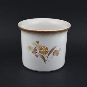 May include: White ceramic container with a floral design featuring orange, yellow, and purple flowers. The rim is brown, and the container has a cylindrical shape. The floral pattern is on the front.