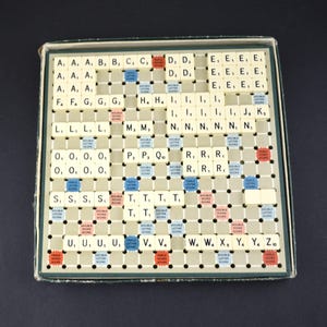 May include: A vintage Scrabble board game, featuring a grid of lettered tiles and colored bonus squares. The game board is housed in a green-edged box, with a dark background. The tiles are beige with black letters and numbers.