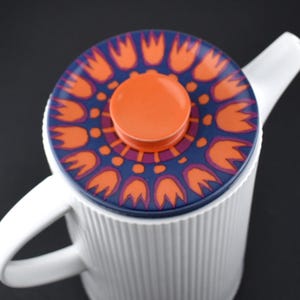 May include: White ceramic teapot with a lid featuring a floral design in orange, purple, and blue. The teapot has a ribbed texture and a curved handle. The lid has an orange knob.